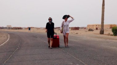 Gözlüklü iki neşeli kız turist ellerinde büyük kırmızı bir bavulla yol boyunca yürüyorlar, otele doğru koşuyorlar.