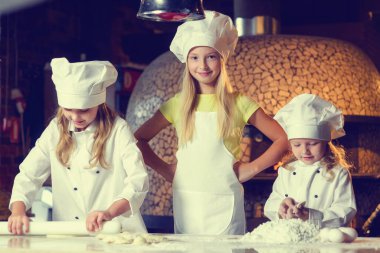 Un içinde, şef üniforması giymiş ve büyük beyaz şapkalar takmış üç neşeli küçük kız bir un ürünü hazırlıyorlar. Yakın plan..