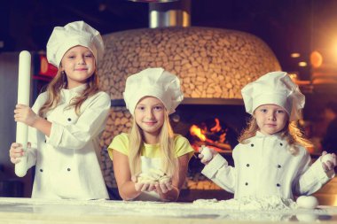 Fırının yanında, un içinde ve aşçı üniforması giymiş üç neşeli küçük kız pizza yapıyorlar..
