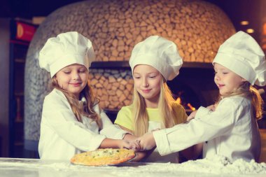 Fırının yanında, un içinde ve aşçı üniforması giymiş üç neşeli küçük kız pizza yapıyorlar..