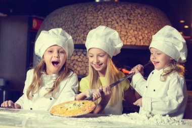 Fırının yanında, un içinde ve aşçı üniforması giymiş üç neşeli küçük kız pizza yapıyorlar..
