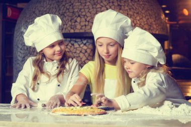 Fırının yanında, un içinde ve aşçı üniforması giymiş üç neşeli küçük kız pizza yapıyorlar..