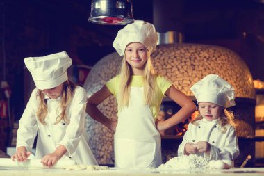 Una bulanmış, şef üniforması giymiş ve büyük beyaz şapkalar takmış üç neşeli küçük kız pizza yapıyor. Yakın plan..