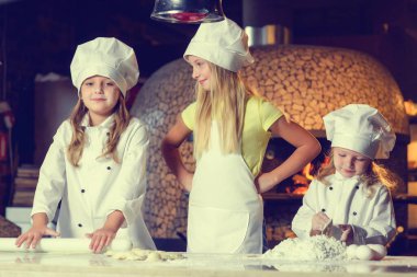 Una bulanmış, şef üniforması giymiş ve büyük beyaz şapkalar takmış üç neşeli küçük kız pizza yapıyor. Yakın plan..