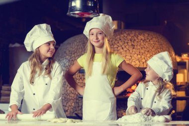 Una bulanmış, şef üniforması giymiş ve büyük beyaz şapkalar takmış üç neşeli küçük kız pizza yapıyor. Yakın plan..