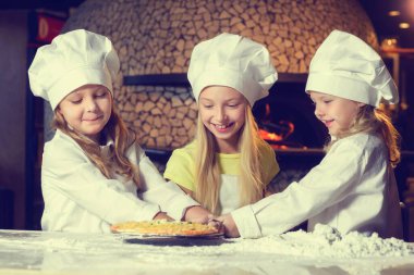 Una bulanmış, şef üniforması giymiş ve büyük beyaz şapkalar takmış üç neşeli küçük kız pizza yapıyor. Yakın plan..