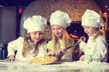 Una bulanmış, şef üniforması giymiş ve büyük beyaz şapkalar takmış üç neşeli küçük kız pizza yapıyor. Yakın plan..