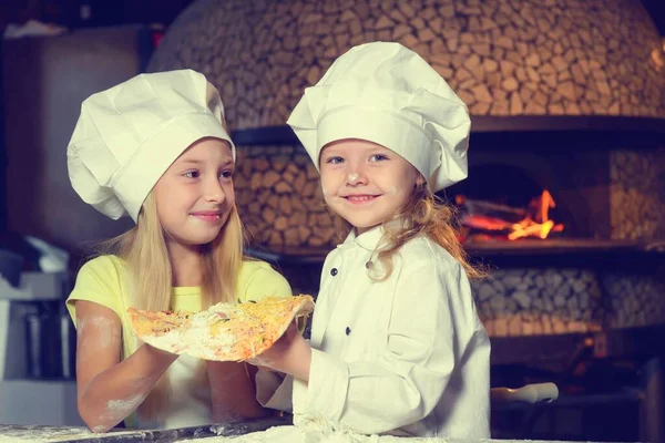 Una bulanmış, şef üniforması giymiş ve büyük beyaz şapkalar takmış iki neşeli küçük kız pizza yapıyorlar. Yakın plan..