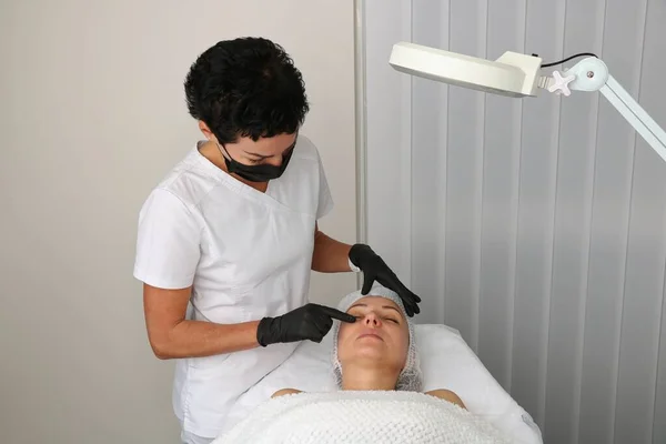 The beautician examines the patient before the facial treatment. — Stock Fotó