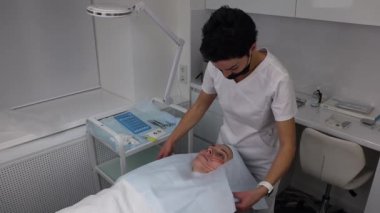 Hasta kanepede yatar, kafası özel bir peçeteyle kaplıdır, kozmetik uzmanı kadını güzellik salonunda yüz gerdirmeye hazırlar..
