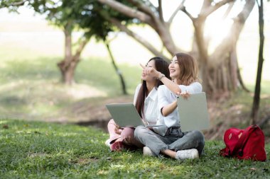 Kampüsün dışında oturan mutlu Asya üniversitesi. İki kız parkta dizüstü bilgisayar ve dijital tabletle birlikte çalışıyor.