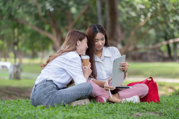 キャンパスの外に座って幸せなアジアの大学を閉じます。公園でノートパソコンやデジタルタブレットと一緒に勉強している2人の女の子が — ストック写真