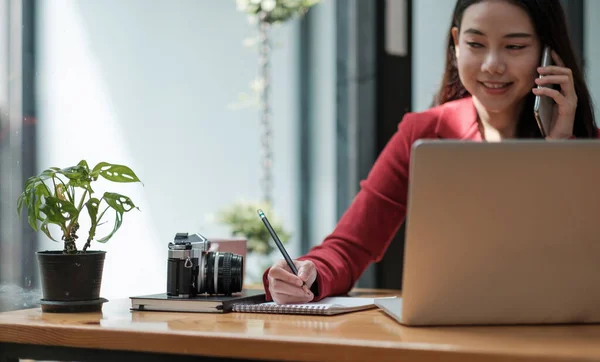Junge Frau telefoniert und schreibt Notizen, während sie an ihrem Schreibtisch sitzt. Asiatischfemale working im home office — Stockfoto