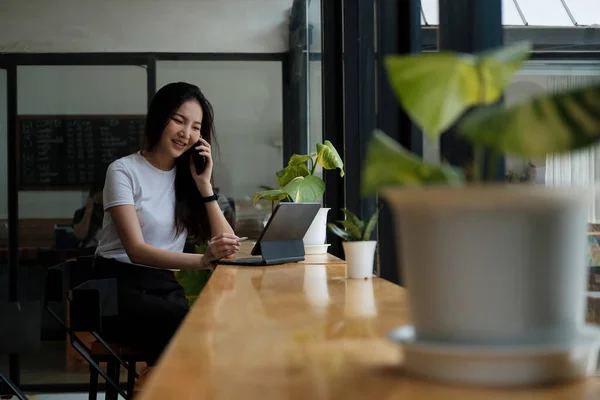 Mujer joven hablando por teléfono móvil y escribiendo notas en una tableta digital mientras está sentada en su escritorio. Asiática mujer trabajando en casa oficina — Foto de Stock