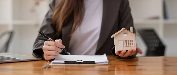 Gros plan agent immobilier femme d'affaires prenant des notes et tenant modèle de maison, assis au bureau avec modèle de maison en papier et les clés, gestionnaire de l'agent immobilier femme, la préparation de documents, hypothèque et la propriété. — Photo