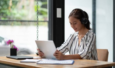 Dinlenme zamanı, kulaklıklı Asyalı kız çalışma alanına oturmuş tabletle müzik dinliyor.