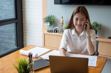 Gülümseyen Asyalı kadın, mali muhasebe numarası, evrak istatistikleri için dizüstü bilgisayarda çalışırken cep telefonuyla konuşuyor.
