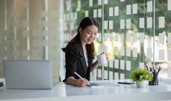 Glückliche schöne junge asiatische Geschäftsfrau, die mit einem digitalen Tablet arbeitet, während sie am Tisch im Co-Working-Büro sitzt. Freiberufliche Tätigkeit, Lächeln, positive Emotionen, Business Financial Concept. — Stockfoto