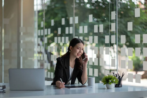 Sonriente joven mujer de negocios asiática sentarse en el escritorio sonrisas hablando en el teléfono inteligente mirando por la ventana disfrutar de una agradable conversación con el cliente, gerente de ventas femenino hace exitoso concepto rentable —  Fotos de Stock