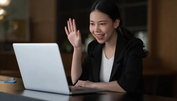 Bild einer glücklichen asiatischen Frau mit weißem Hemd, die lächelt und mit der Hand am Laptop wedelt, während sie im Büro telefoniert oder plaudert. — Stockfoto