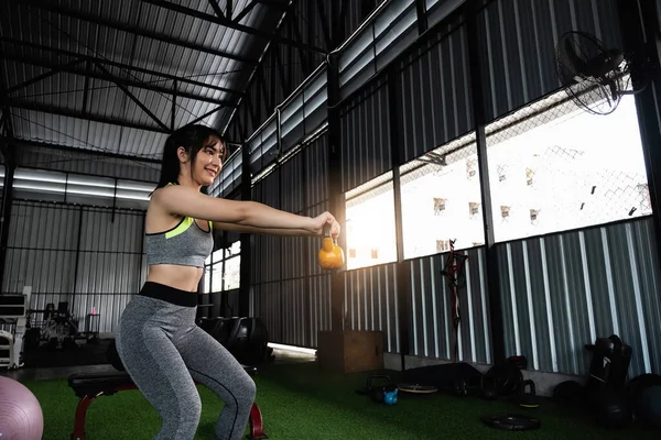 Νεαρή Ασιάτισσα απολαμβάνει την άσκηση με την ανύψωση καμπάνα βραστήρα με το ένα χέρι στο γυμναστήριο. έννοια bodybuilding αθλητισμού. — Φωτογραφία Αρχείου