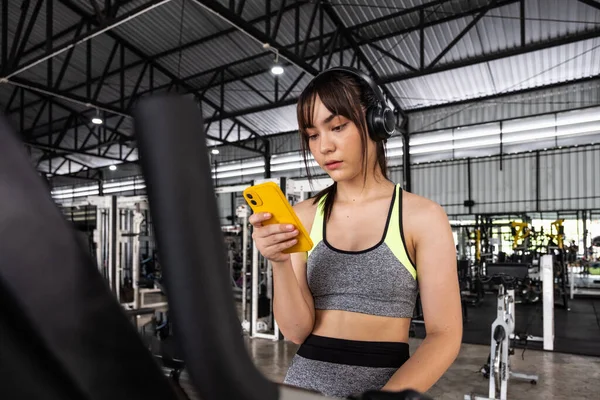 Joven asiática feliz mujer sentada y escuchando música por teléfono inteligente mientras hace ejercicio en el gimnasio — Foto de Stock