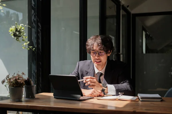 Asiático joven hombre de negocios en gafas de trabajo con la tableta digital de forma remota, sentado en la mesa de madera en el café. Agradable hombre feliz comunicarse en la red social, la búsqueda de información en línea — Foto de Stock