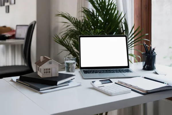 Bärbar dator med tom vit skärm på modernt skrivbord, fastighet med husmodell, anteckningsblock och miniräknare på arbetsplatsen — Stockfoto