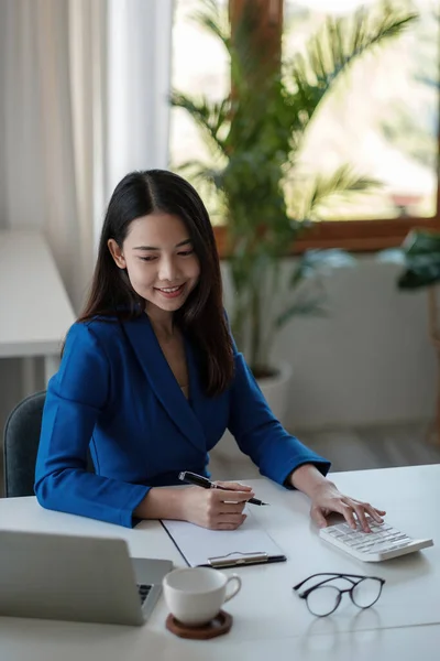 Πορτρέτο νεαρή Ασιάτισσα επιχειρηματίας κάθεται στο γραφείο τους και να κρατήσει σημειώσεις χρησιμοποιώντας φορητό υπολογιστή και αριθμομηχανή στο γραφείο. — Φωτογραφία Αρχείου