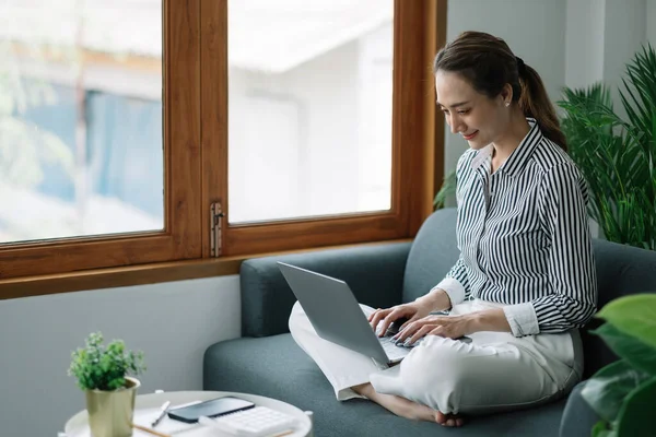 Ασιάτισσα νεαρή γυναίκα που εργάζεται από το γραφείο στο σπίτι κάθεται στον καναπέ κρατήστε το χαρτί τιμολόγιο ελέγξετε πληροφορίες online — Φωτογραφία Αρχείου