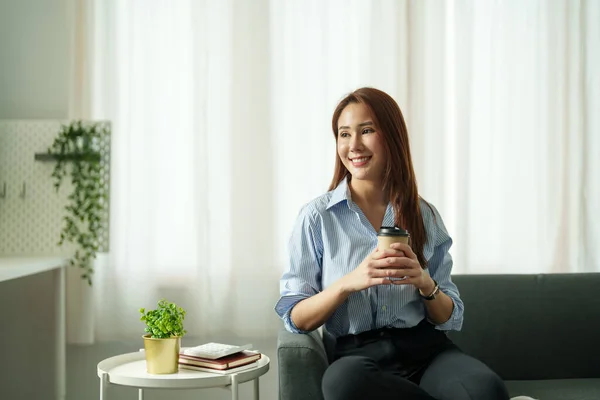 Gelukkig aantrekkelijke aziatische vrouw hand holding kopje hete koffie in de woonkamer op zoek naar buiten. — Stockfoto