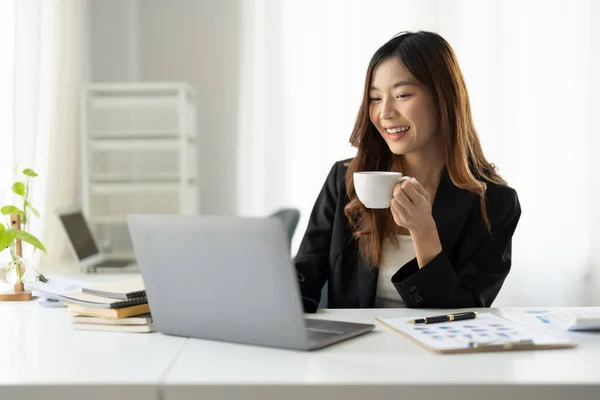Mutlu Asyalı iş kadını sabahları dizüstü bilgisayarla çalışırken elinde bir fincan kahve tutuyor. — Stok fotoğraf