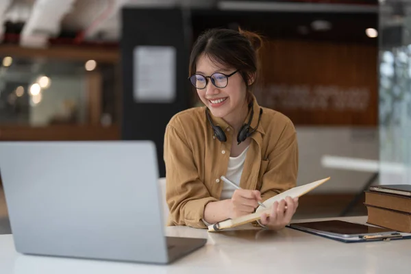 Portrait de jeune femme asiatique à l'aide d'un ordinateur portable et l'écriture de liste de prise de notes dans le bloc-notes de travail ou d'apprentissage sur ordinateur portable à l'intérieur- cours éducatif ou de formation, séminaire, concept d'éducation en ligne. — Photo