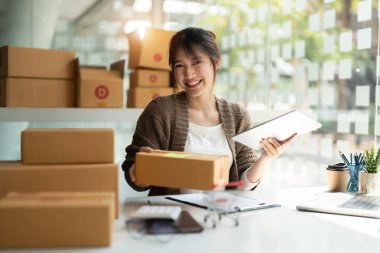 Genç bayan küçük işletme sahibi evden online alışveriş yapıyor. Arka planda dijital tabletle paket ürünü hazırlayan müşterilerin siparişlerini kontrol ediyorum. KOBİ girişimcisi veya serbest meslek kavramı