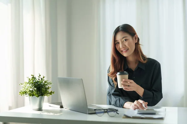 Çekici Asyalı kadın ofis çalışanı dizüstü bilgisayarda çalışıyor ve modern bir ofiste kahve içmekten zevk alıyor.. — Stok fotoğraf