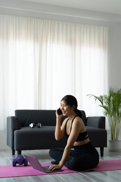 Portret azjatyckiej sportowej kobiety w odzieży sportowej ćwiczącej i korzystającej z laptopa i telefonu w domu w salonie, siedzącej na podłodze z hantlami na macie do jogi. Sport i koncepcja szkolenia online — Zdjęcie stockowe