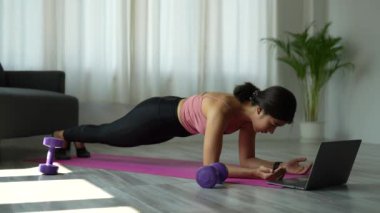 Güzel Asyalı fitness kadını bir tahta egzersizi yapıyor dizüstü bilgisayarda ders izliyor, oturma odasında antrenman yapıyor. Sağlıklı yaşam tarzı