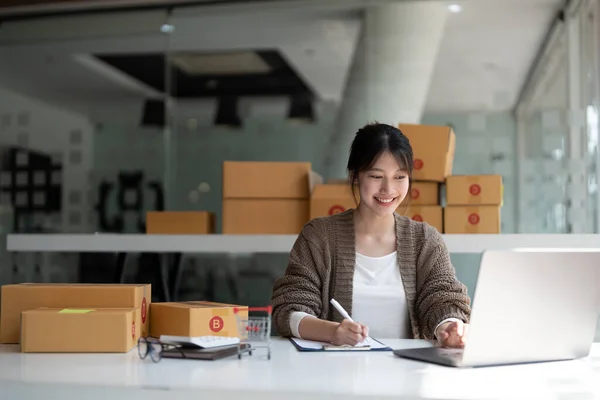 Eladó online, Fiatal ázsiai nő ellenőrzése érdekében, és az ügyfelek adatait és címét a laptop vagy a doboz annak érdekében, hogy felkészüljenek a szállítás szerint az információ, Online vásárlás kkv-vállalkozó — Stock Fotó