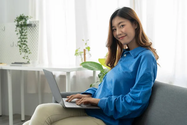 Asyalı genç iş kadını evde dizüstü bilgisayarla gülüyor ve çalışıyor oturma odasındaki kanepede internetten internete bağlanıyor, serbest çalışan kız kanepeye dizüstü bilgisayar kullanıyor, rahat, yeni normal yaşam tarzı konsepti kullanıyor. — Stok fotoğraf