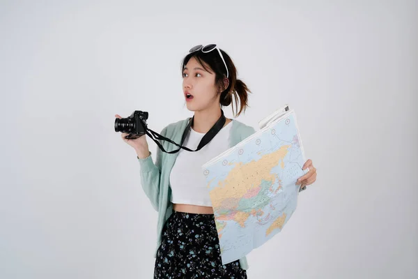 Portrait of smiling young female Asian tourist backpacker holding map and camera in isolated white background - summer travel concept — стокове фото