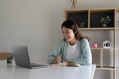 Asyalı genç bir kadın not defterine not alıyor. Kapalı alanda çalışıyor veya öğreniyor. Eğitim kursu, seminer, eğitim, çevrimiçi eğitim konsepti.