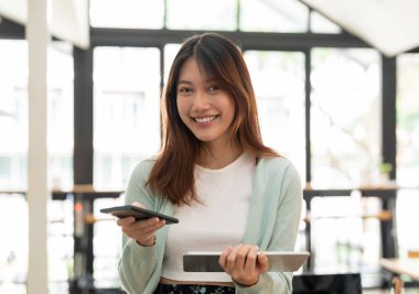 Kamerada cep telefonu ve dijital tablet kullanan Asyalı genç bir kadın görülüyor.