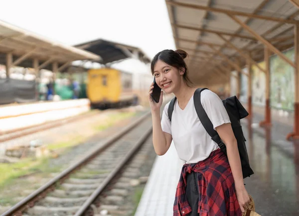 年轻美丽的亚洲女人，背着背包，站在站台上的火车旁边，一边用电话。便宜旅游暑期的概念 — 图库照片