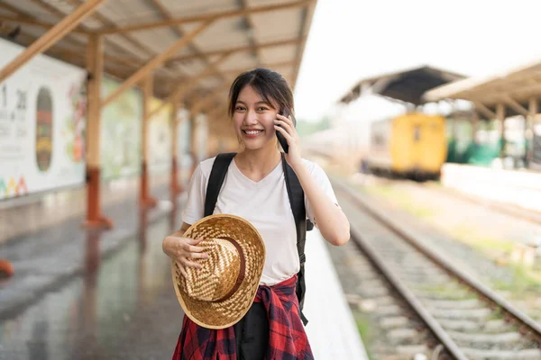 Sırt çantalı genç Asyalı güzel bir kadın platformda demiryolu treninin yanında dururken telefonu kullanıyor. Ucuz seyahat yaz konsepti — Stok fotoğraf