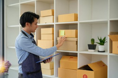 Genç, çekici Asyalı girişimci kamera işine bakın. Evdeki kutudan mutlu bir şekilde bakın. Paket teslimatını sme tedarik zinciri, tedarik, omnichannel veya koronavirüs karantina konseptine hazırlayın.