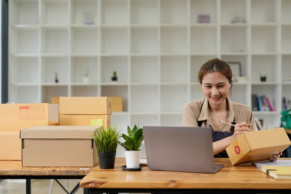 Young smiling beautiful owner asian woman freelance sme business online shopping working on laptop computer with parcel box on desk at home - SME business online and delivery concept — Stock Fotó