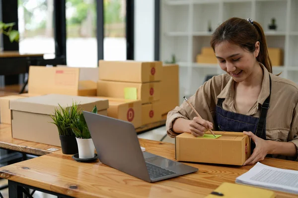 Genç gülümseyen güzel sahibi Asyalı kadın serbest meslek edinmiş dizüstü bilgisayarla evde paket kutusuyla alışveriş yapıyor - KOBİ işi online ve teslimat kavramı
