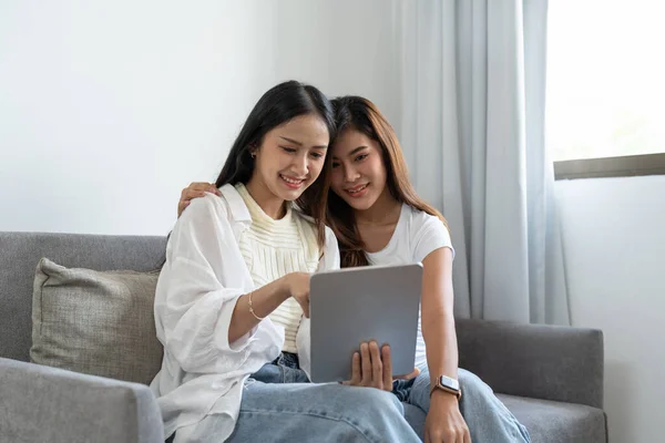 Gelukkig jong aziatisch lesbisch paar knuffelen plezier met behulp van digitale tablet ontspannen op de bank thuis. Twee lachende vrouwen vrienden houden computer kijken naar het scherm genieten van surfen online kijken video — Stockfoto