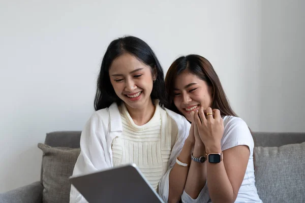 快乐的年轻的亚洲女同性恋夫妇拥抱有乐趣使用数字平板电脑放松在沙发上的家。两位微笑的女性朋友抱着电脑看屏幕，享受上网观看视频 — 图库照片