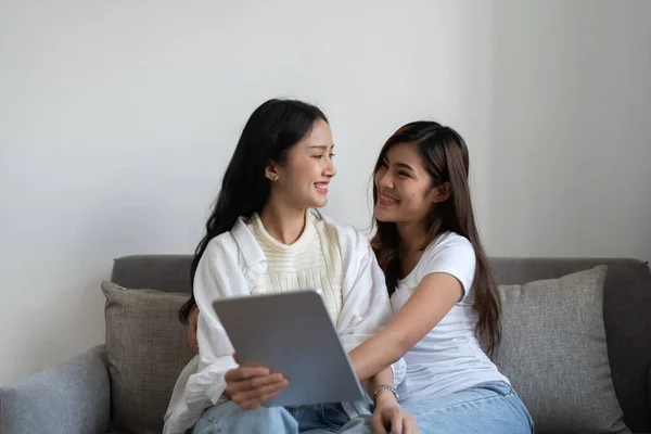 Feliz joven asiática lesbiana pareja abrazo tener divertido usando digital tablet relajarse en sofá en casa. Dos amigas sonrientes sosteniendo la computadora mirando la pantalla disfrutando de navegar online viendo video —  Fotos de Stock